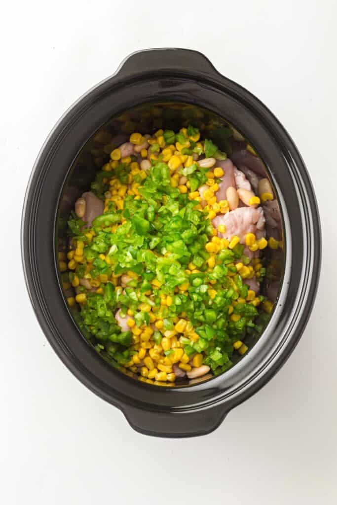 A slow cooker filled with the ingredients for Slow Cooker White Chicken Chili&mdash;raw chicken, corn, white beans, and chopped green peppers&mdash;sits on a white background.