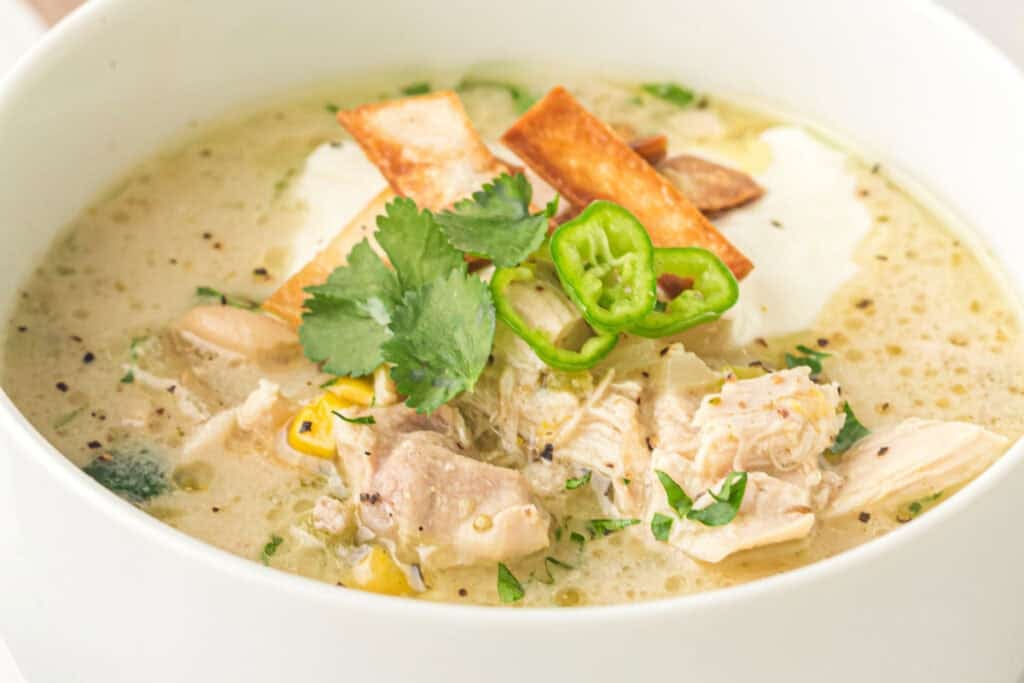 A bowl of creamy Slow Cooker White Chicken Chili garnished with cilantro, sliced green pepper, tortilla strips, and a dollop of sour cream.