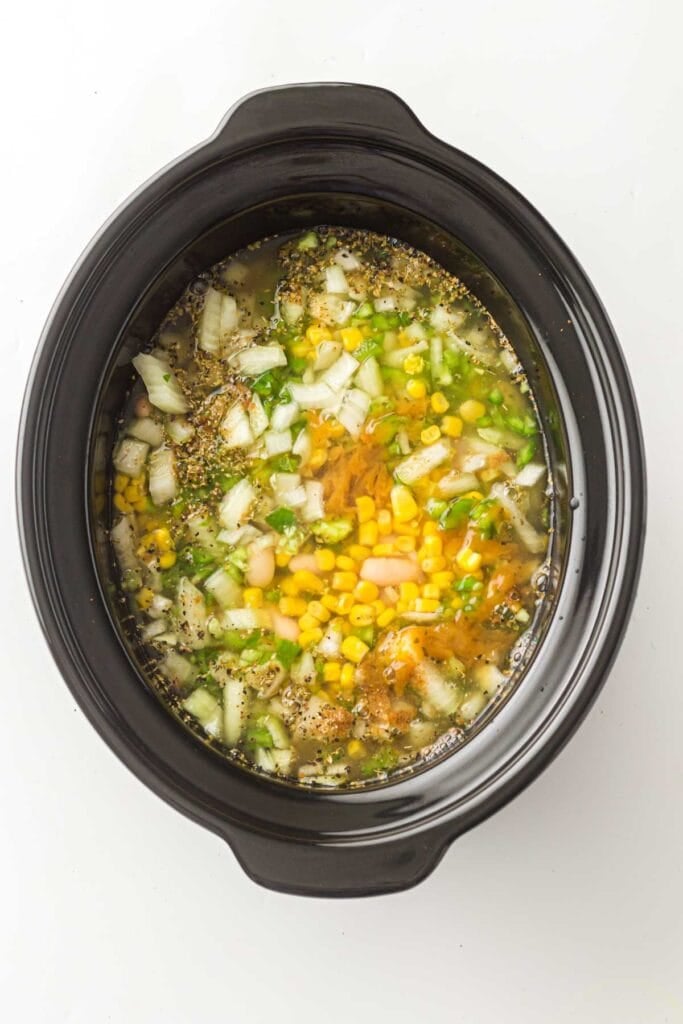A slow cooker filled with uncooked Slow Cooker White Chicken Chili ingredients, including chopped onions, corn, white beans, shredded carrots, green peppers, and spices in broth.