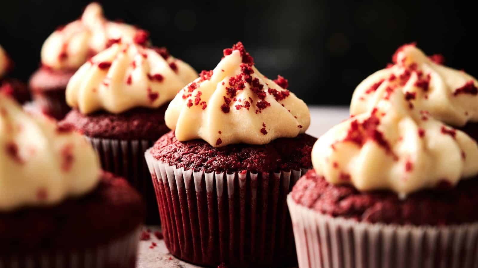 Close-up of several red velvet cupcakes topped with cream cheese frosting and red cake crumbs, arranged in rows on a surface.