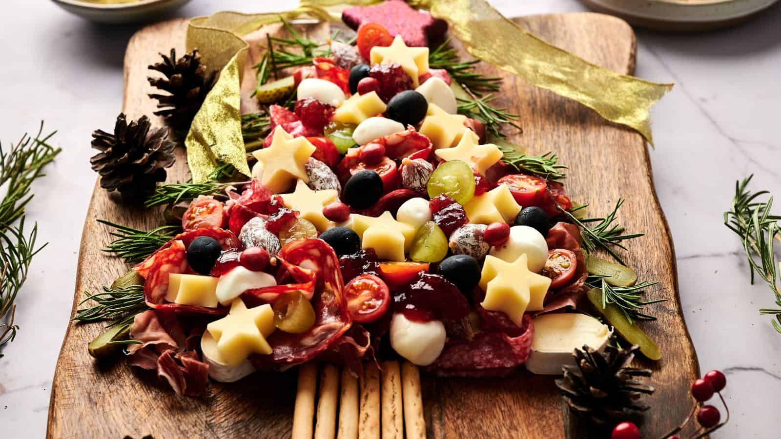 A charcuterie board arranged in the shape of a Christmas tree with cheese stars, meats, olives, tomatoes, grapes, and breadsticks on a wooden board, garnished with rosemary.