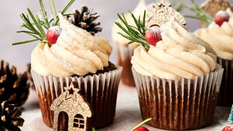 Cupcakes with swirled frosting are decorated with cranberries, rosemary, powdered sugar, pinecones, and gold holiday-themed toppers.