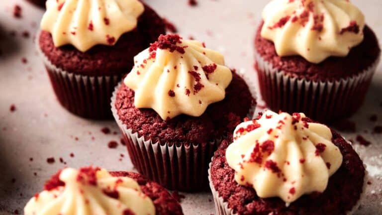 Red velvet cupcakes with cream cheese frosting and red crumbs are arranged on a light surface.