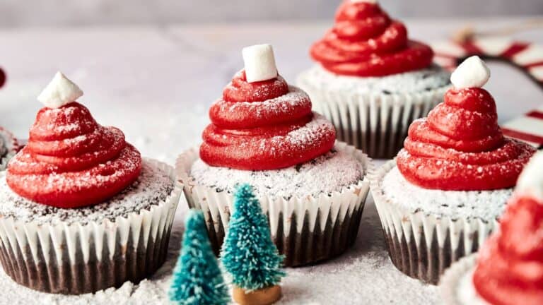Chocolate cupcakes topped with red frosting shaped like Santa hats, dusted with powdered sugar, each finished with a small marshmallow on top. Miniature tree decorations in foreground.