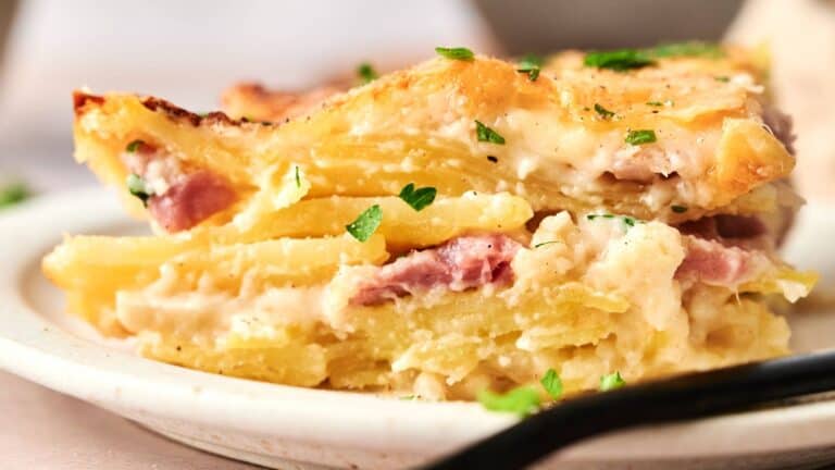 A close-up of a slice of potato gratin with ham and creamy sauce, garnished with chopped parsley, served on a white plate.