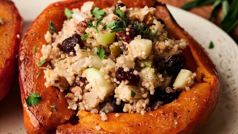 Baked acorn squash half filled with a mixture of grains, chopped apples, nuts, dried fruit, and fresh herbs on a white plate.