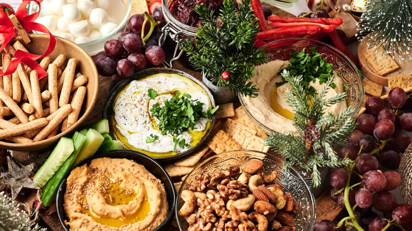 A holiday-themed platter with bowls of hummus, yogurt dip, nuts, grapes, breadsticks, and crackers, decorated with festive greenery and red ribbons.