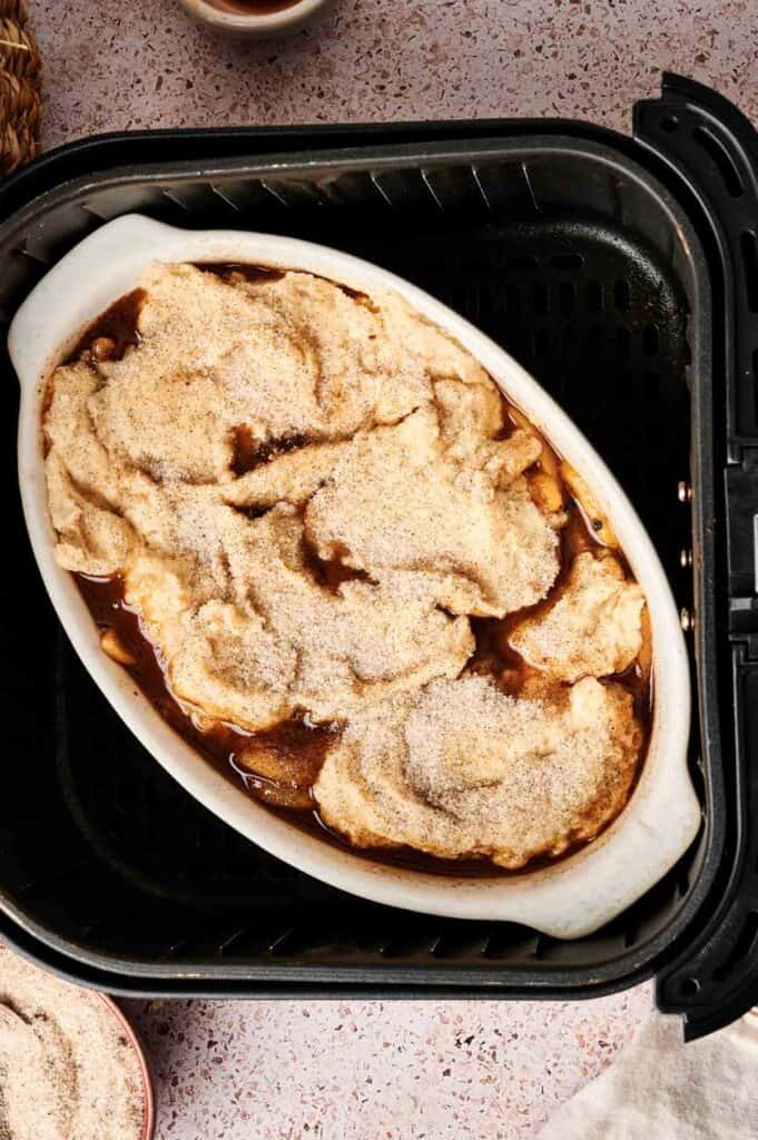 An oval baking dish filled with unbaked Air Fryer Apple Cobbler, topped with dough and cinnamon sugar, sits inside an air fryer basket.