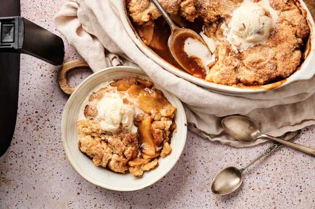 A bowl of Air Fryer Apple Cobbler topped with vanilla ice cream sits beside a larger baking dish of cobbler, with serving spoons and two empty spoons on a light countertop.