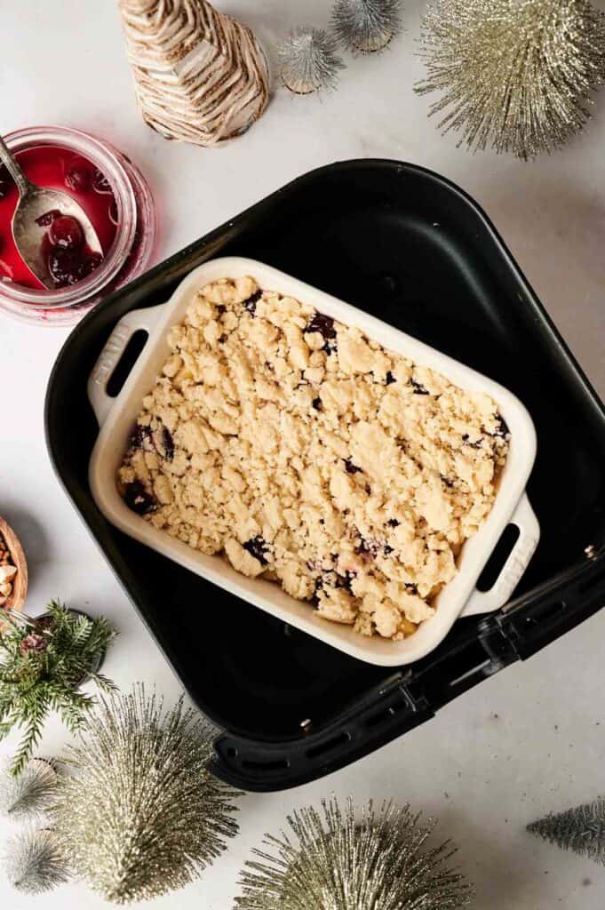 A rectangular baking dish with an unbaked Air Fryer Christmas Dump Cake sits inside an air fryer basket, surrounded by holiday decorations and a jar of cherries.