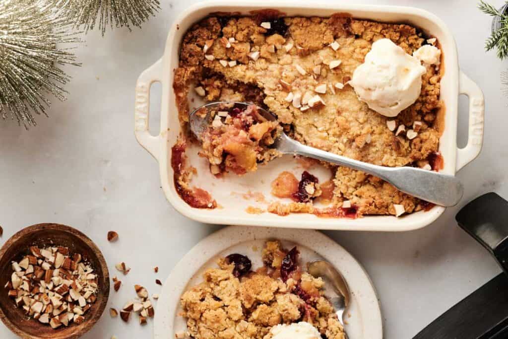 Baked fruit crumble, similar to an Air Fryer Christmas Dump Cake, topped with ice cream in a ceramic dish, with a serving spoon, chopped nuts in a bowl, and a portion on a plate beside it.