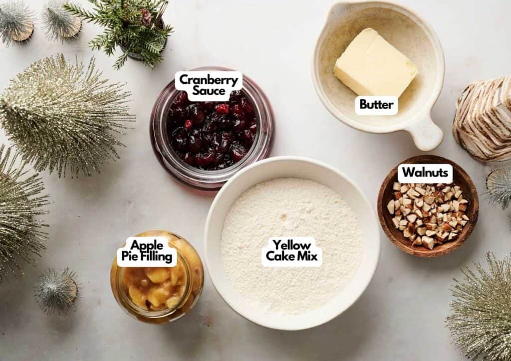Five ingredients on a white surface for an Air Fryer Christmas Dump Cake: cranberry sauce, butter, walnuts, apple pie filling, and yellow cake mix in bowls, with festive holiday tree decorations nearby.