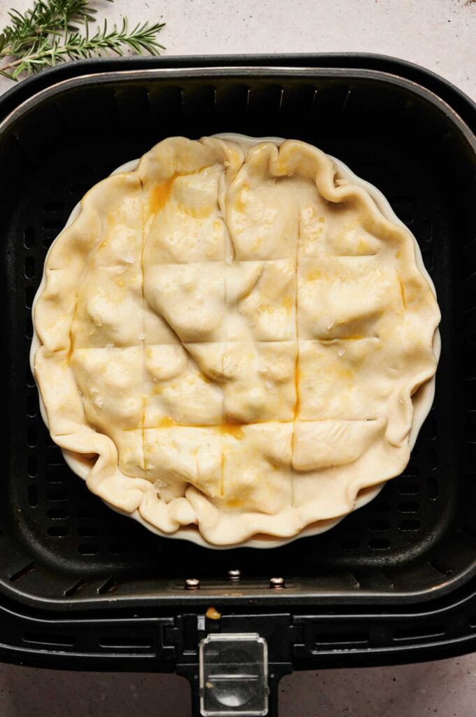 Unbaked Air Fryer Turkey Pot Pie with a crimped crust and vent slits sits in an air fryer basket, ready to be cooked. A sprig of rosemary is visible in the corner.