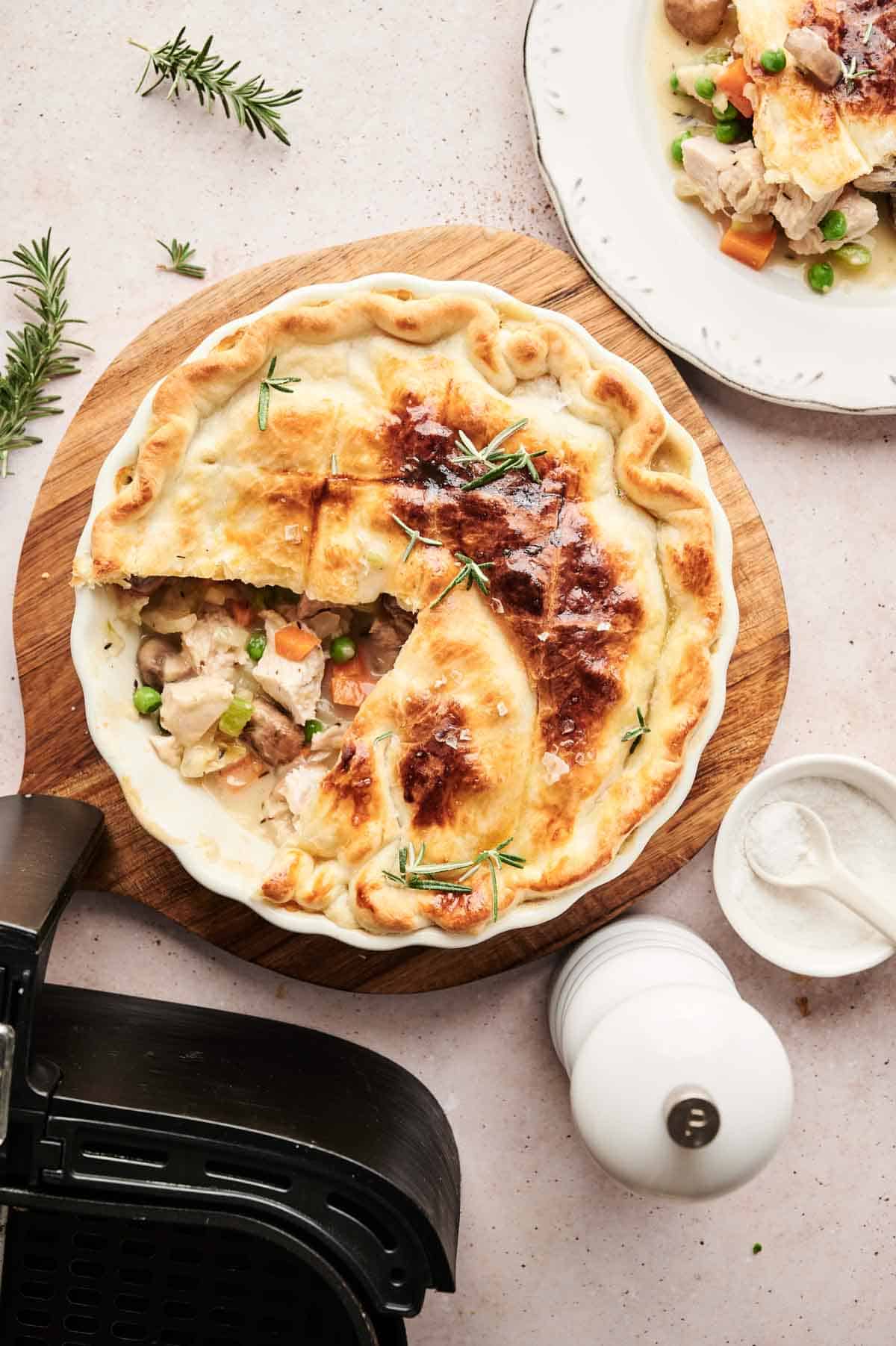 A baked chicken pot pie with a golden crust sits on a wooden board, partially sliced to reveal the filling, while a serving of air fryer turkey pot pie is plated nearby for a delicious comparison.
