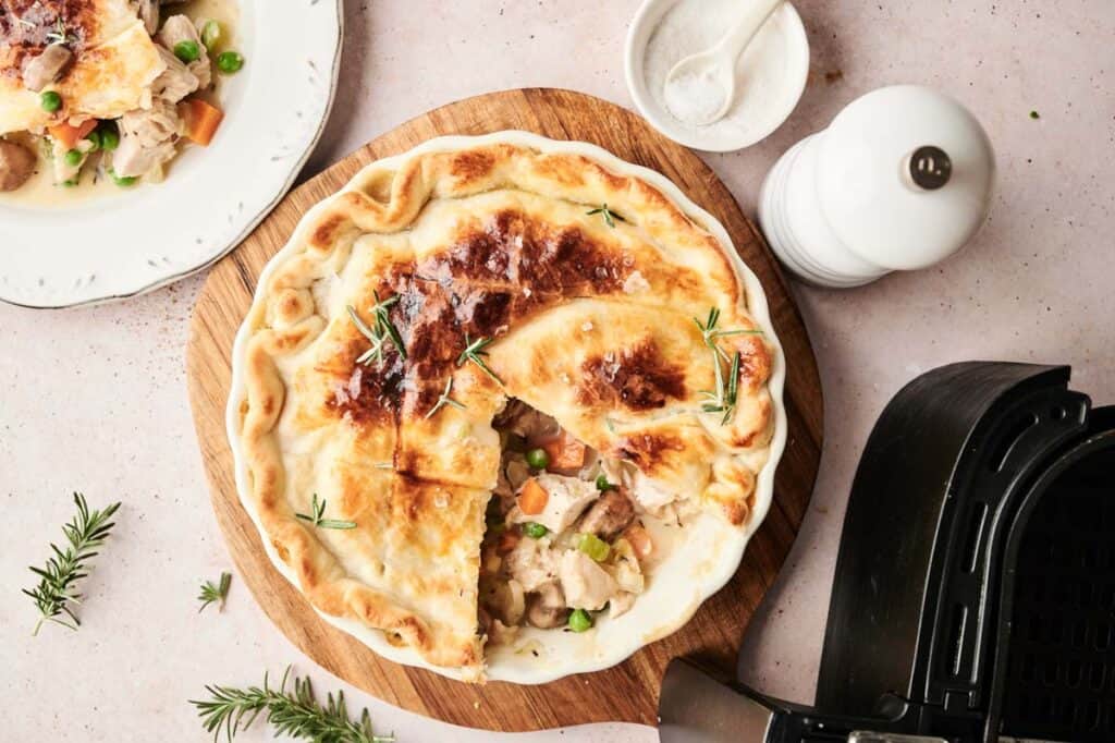 A golden-brown Air Fryer Turkey Pot Pie with a slice removed reveals a hearty filling of turkey and vegetables, sitting on a wooden board next to an air fryer and a plate with a generous serving.