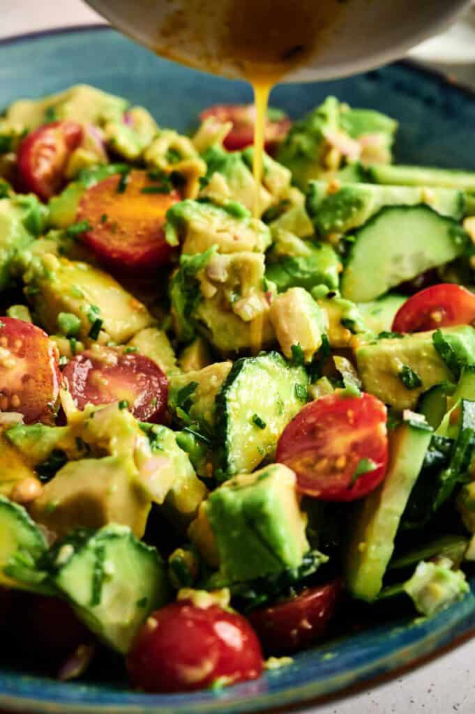 A close-up of a fresh Avocado Salad with cherry tomatoes, cucumber, and herbs, as dressing is poured on top.
