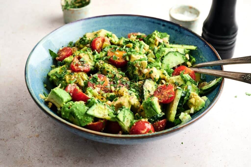 A blue bowl filled with a fresh Avocado Salad of cherry tomatoes, cucumber, and herbs, with salad servers resting inside.