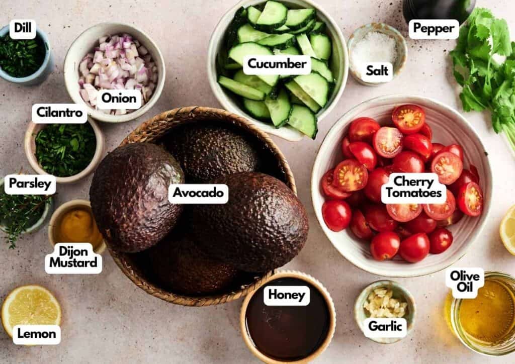Assorted fresh ingredients for an Avocado Salad, including avocado, cherry tomatoes, cucumber, onion, herbs, lemon, garlic, honey, dijon mustard, olive oil, salt, and pepper arranged in bowls on a table.