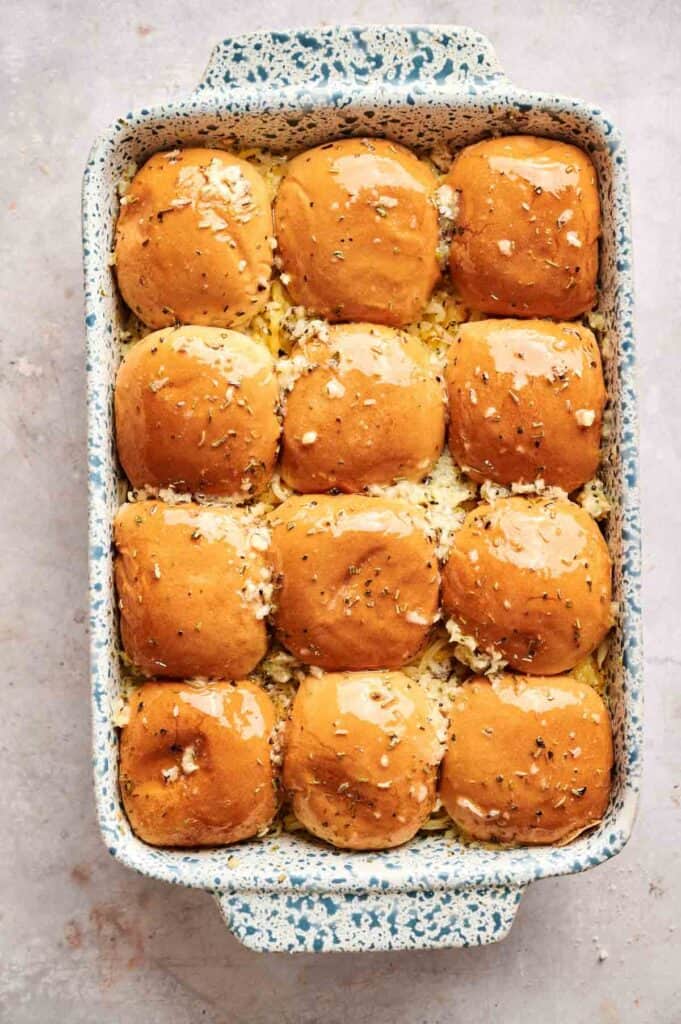 A baking dish filled with twelve golden-brown dinner rolls, reminiscent of Buffalo Chicken Sliders, topped with herbs and melted butter.