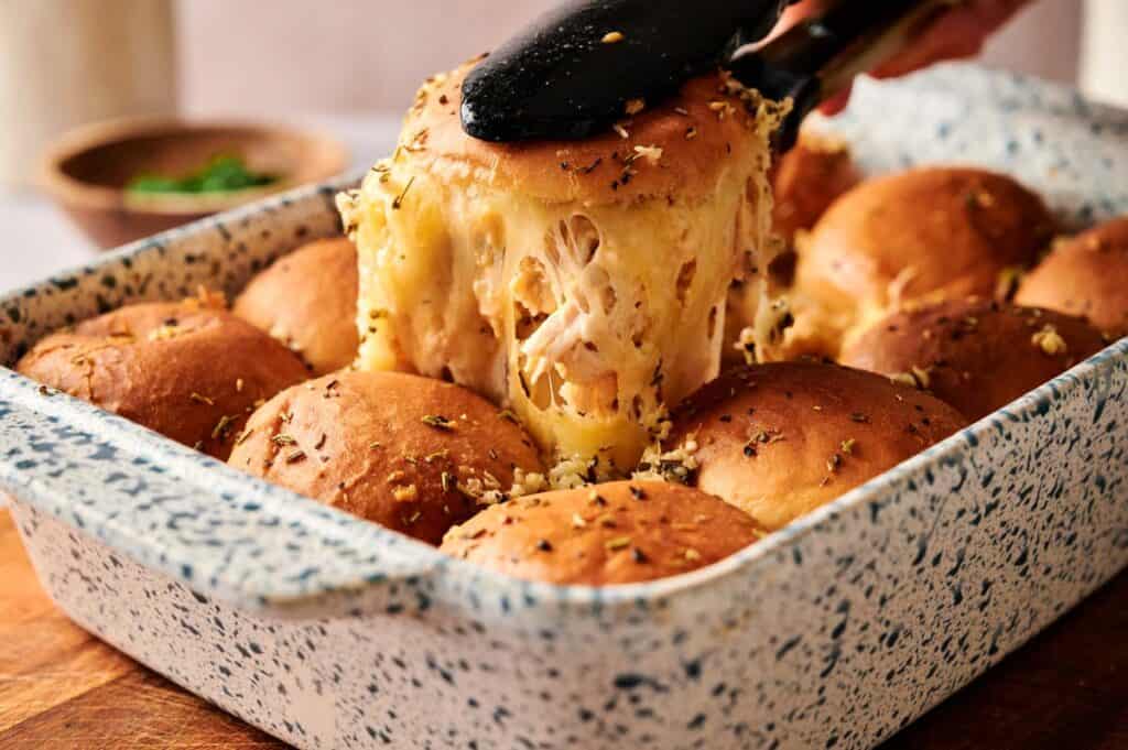 A hand uses tongs to lift a cheesy Buffalo Chicken Slider bun from a baking dish filled with golden brown rolls sprinkled with herbs.