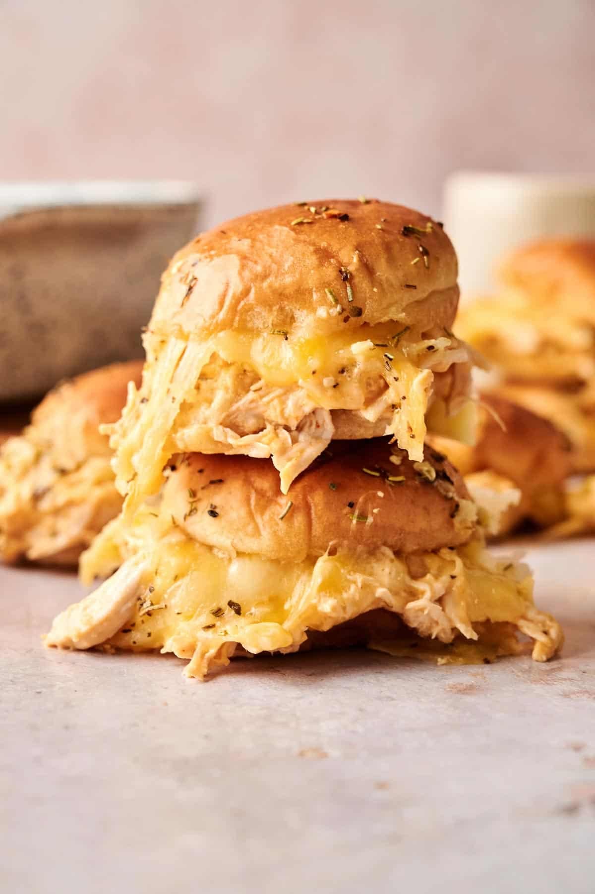 Two stacked Buffalo Chicken Sliders on golden buns with shredded chicken and melted cheese spilling out, set on a light surface with more sliders in the background.