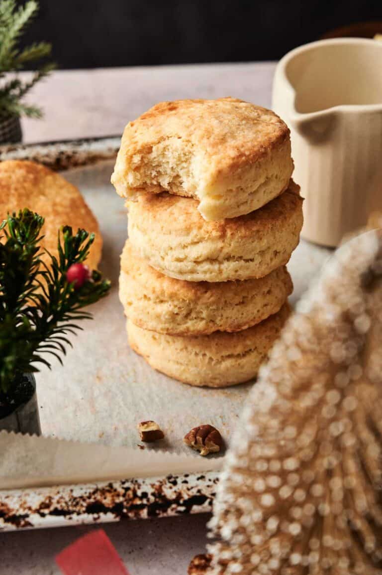 A stack of three golden buttermilk biscuits on a baking tray, with the top biscuit partially bitten, sits next to a small plant and a cream-colored pitcher.