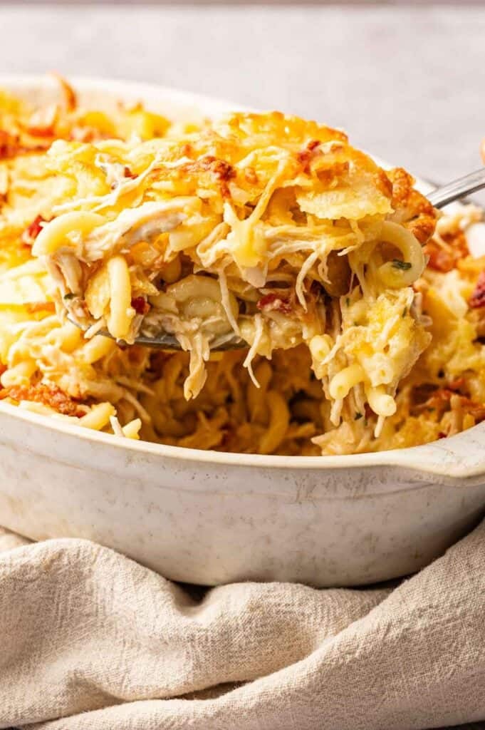 A serving of Chicken Bacon Ranch Casserole with shredded chicken, melted cheese, and crispy bacon is being lifted from a white baking dish.
