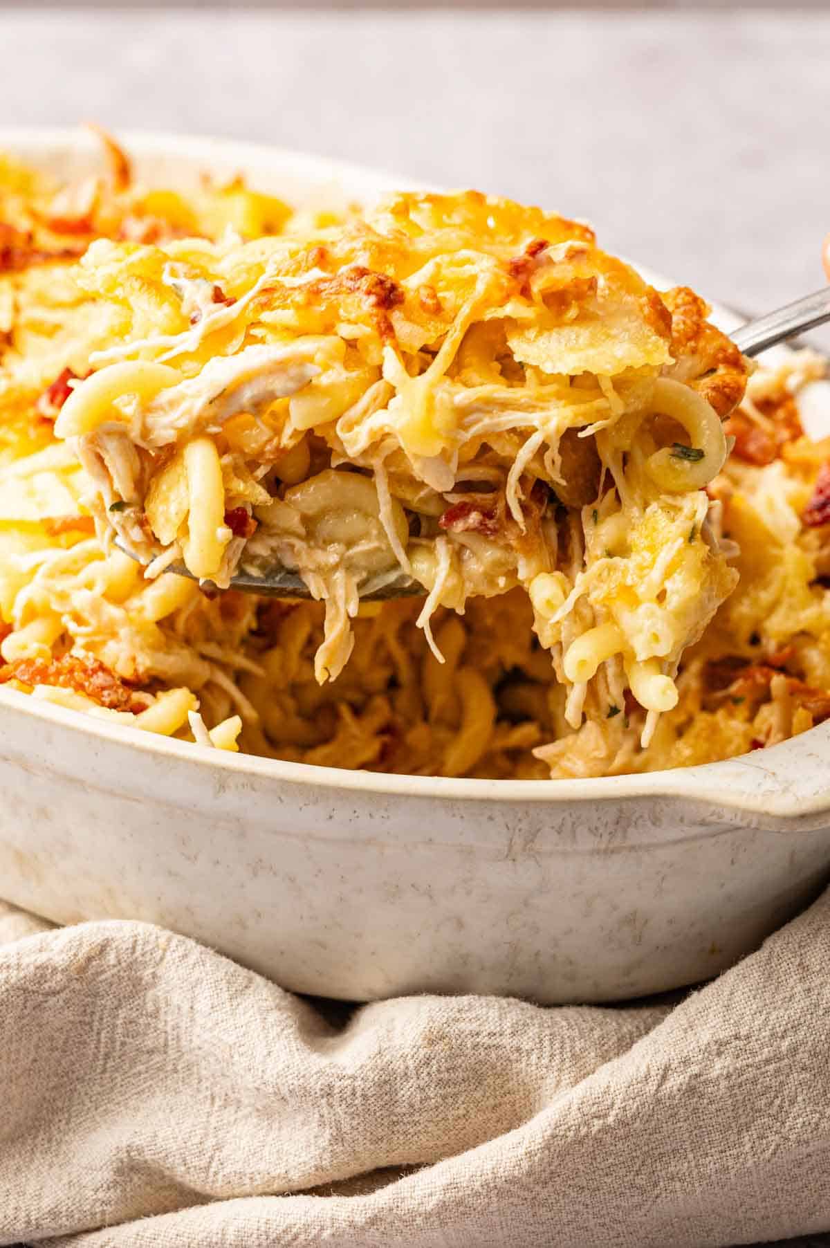 A serving of Chicken Bacon Ranch Casserole with shredded chicken, melted cheese, and crispy bacon is being lifted from a white baking dish.