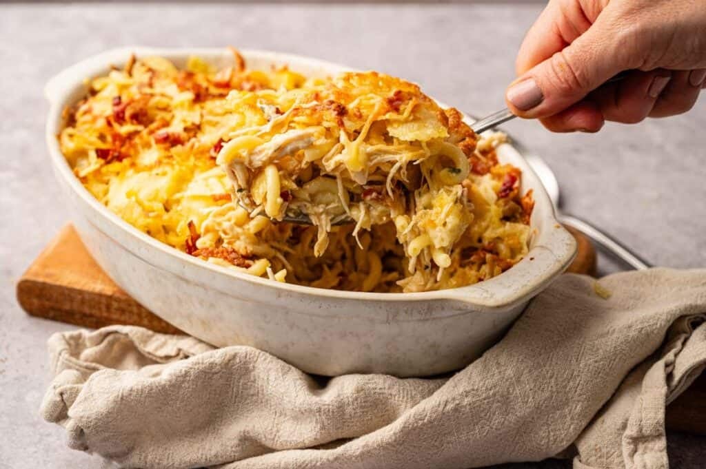 A hand serves a spoonful of Chicken Bacon Ranch Casserole, topped with melted cheese and crispy toppings, from a white oval dish.