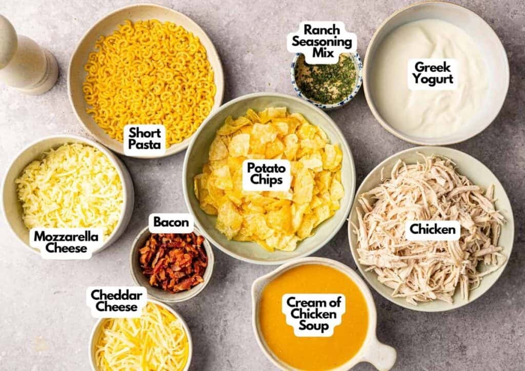 A top-down view of bowls with labeled ingredients for Chicken Bacon Ranch Casserole: short pasta, mozzarella cheese, cheddar cheese, bacon, potato chips, cream of chicken soup, chicken, ranch seasoning mix, and Greek yogurt.