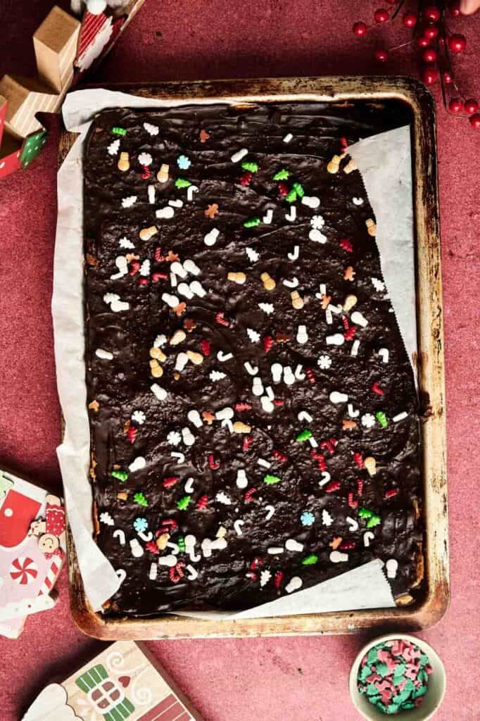 A baking tray lined with parchment paper holds Christmas Crack fudge topped with colorful holiday-themed sprinkles on a red surface.