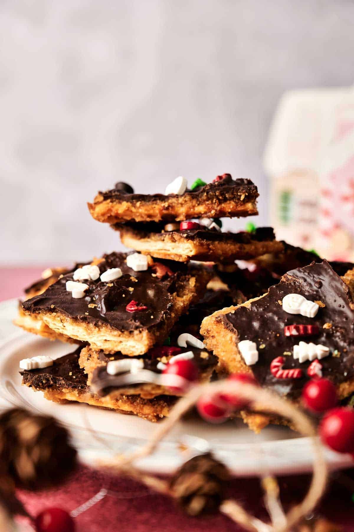 A plate of chocolate-topped Christmas Crack cookie bars, decorated with festive holiday sprinkles, stacked on a white dish.