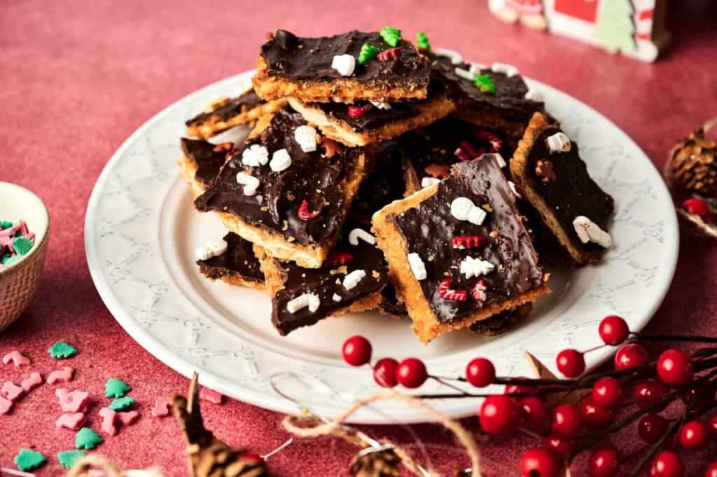 A plate of Christmas Crack dessert bars, covered in chocolate and topped with festive holiday sprinkles, sits on a pink surface with seasonal decorations nearby.
