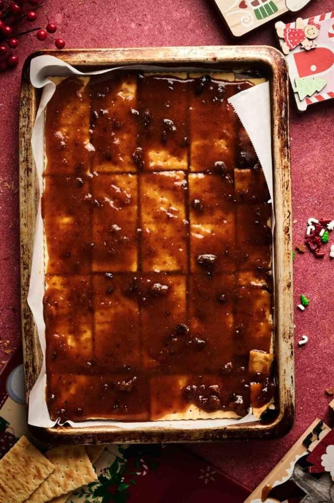 A baking tray lined with parchment and topped with square crackers covered in a glossy brown sauce, resembling Christmas Crack, sits on a red surface with holiday-themed cards and decorations nearby.