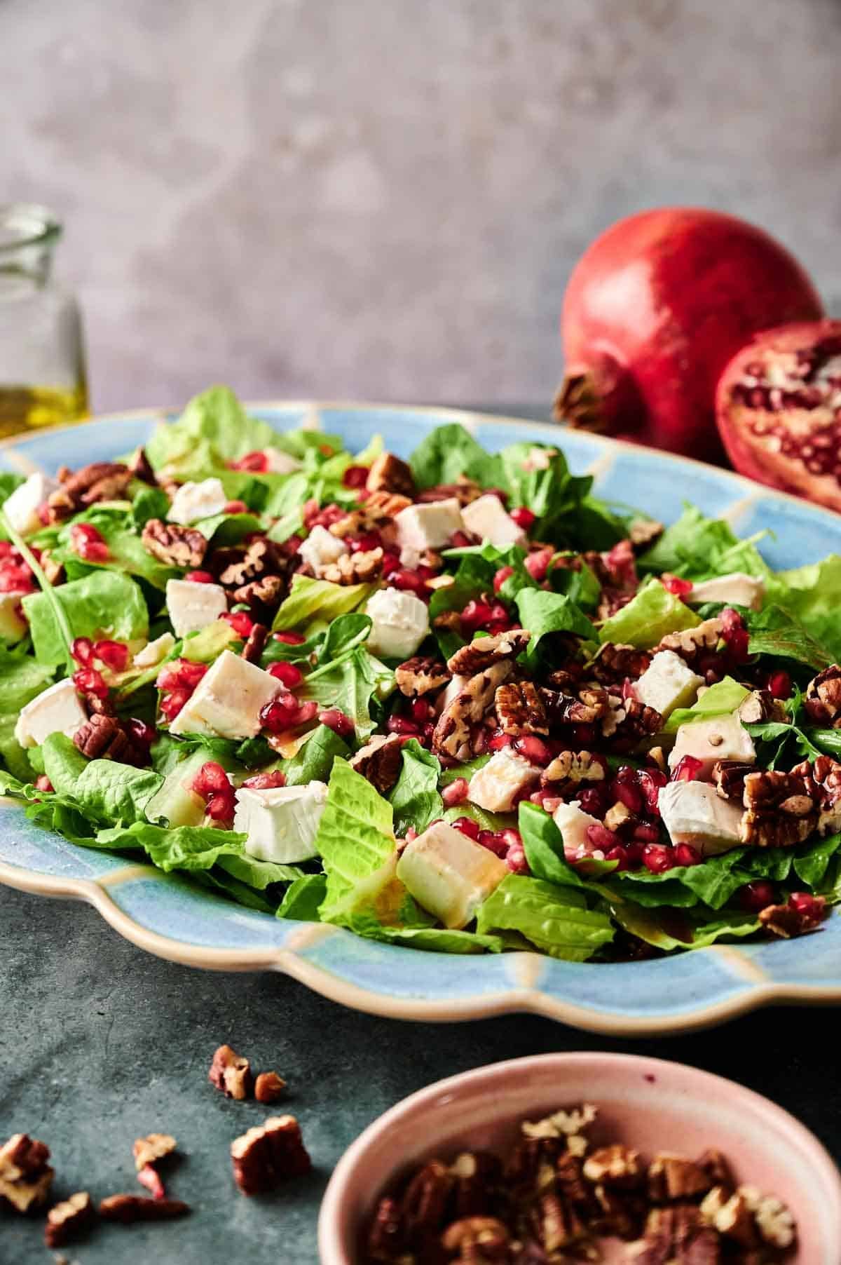 A plate of Christmas Salad with romaine lettuce, feta cheese cubes, pomegranate seeds, and chopped pecans, with a festive pomegranate and a small bowl of pecans in the background.