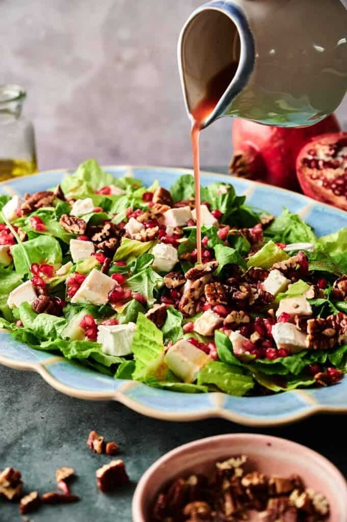 A plate of Christmas Salad with lettuce, pomegranate seeds, cheese cubes, and pecans is being drizzled with vinaigrette from a small pitcher.
