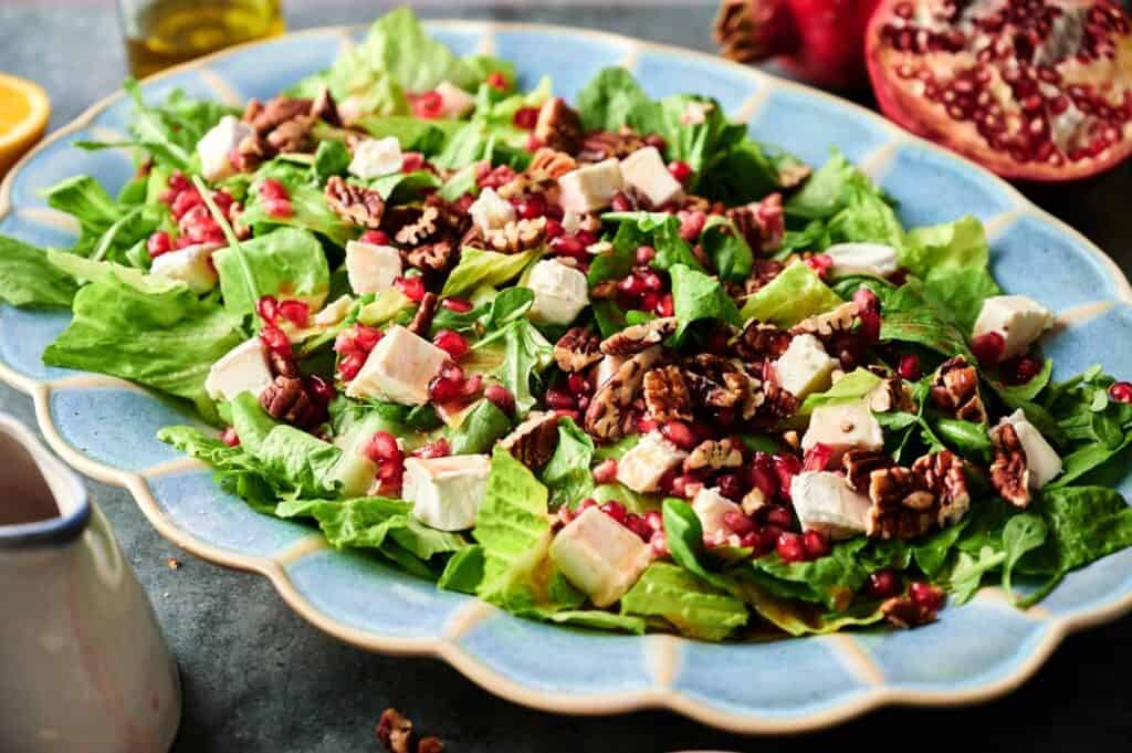 A festive Christmas Salad featuring romaine lettuce, feta cheese cubes, pecans, and pomegranate seeds is artfully arranged on a blue oval plate.