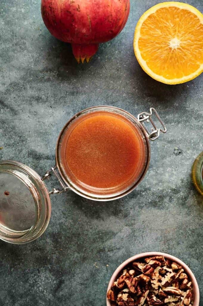 A glass jar of brown liquid dressing with an open lid sits on a gray surface, surrounded by a pomegranate, a halved orange, and a bowl of pecans&mdash;perfect ingredients for a festive Christmas Salad.