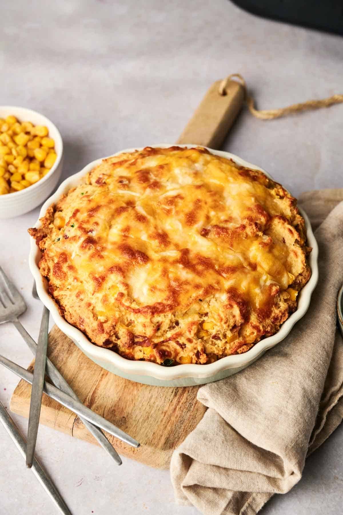 An Air Fryer Cornbread Casserole, golden and cheesy, sits in a round dish on a wooden board beside a beige napkin, utensils, and a small bowl of corn.