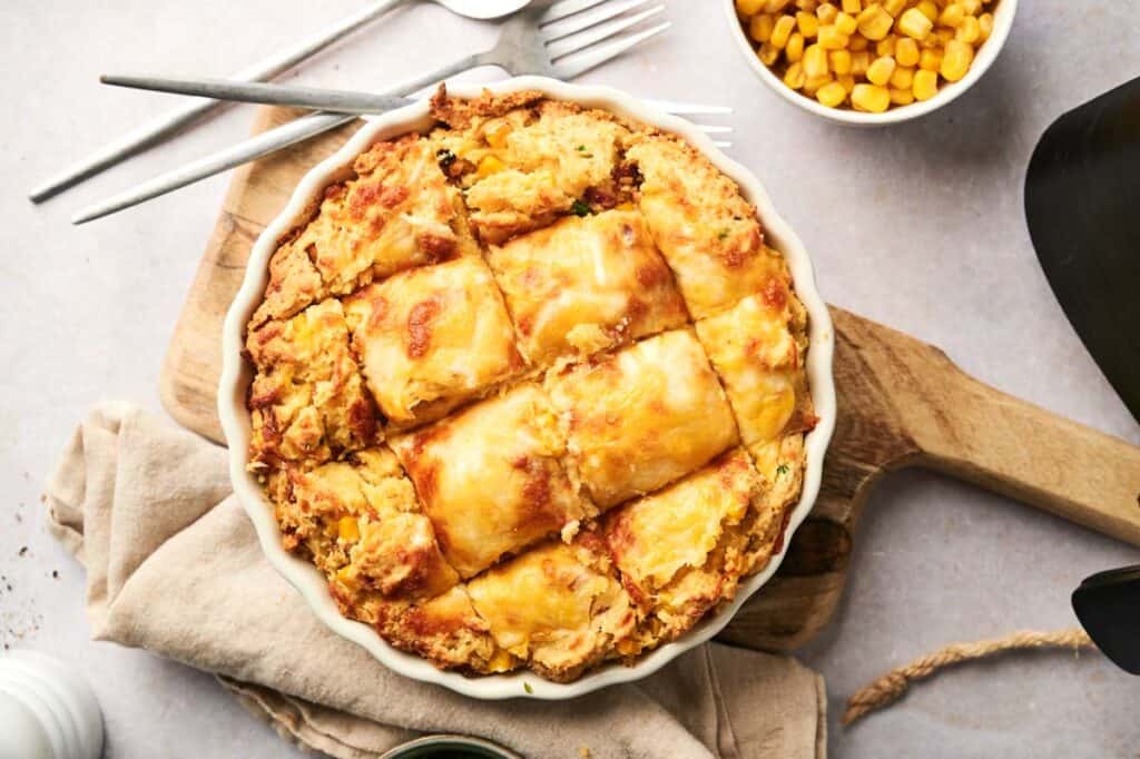 A golden Air Fryer Cornbread Casserole with a flaky crust is cut into six squares, served on a wooden board alongside a napkin, forks, and a bowl of fresh corn.