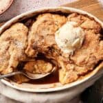 An Air Fryer Apple Cobbler in a white dish with a missing scoop, topped with vanilla ice cream and served with a spoon inside.