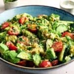 A blue bowl filled with a fresh Avocado Salad, featuring cherry tomatoes, cucumber, chopped herbs, and a sprinkle of ground pepper.