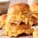 A close-up of two stacked Buffalo Chicken Sliders and cheese sliders on soft buns, with melted cheese and herbs visible. More sliders are blurred in the background.