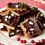 A plate of Christmas Crack—chocolate-covered toffee squares topped with festive holiday-themed sprinkles—rests on a white plate with red decorations nearby.