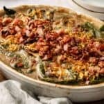 Green Bean and Bacon Casserole topped with chopped bacon, crispy fried onions, and leafy greens in a white oval dish.