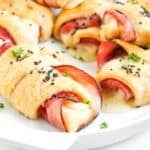 Golden Ham and Cheese Crescent Rolls filled with savory ham and melted cheese, topped with black sesame seeds and garnished with chopped parsley, served on a white plate.