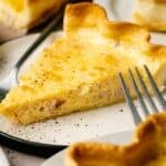 A slice of Ham and Cheese Quiche with a golden crust sits on a plate, garnished with black pepper, with a fork beside it.