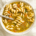 A bowl of spiral pasta soup with chunks of chicken, carrots, celery, and herbs—just like a comforting Instant Pot Chicken Noodle Soup—with a spoon resting inside the bowl.