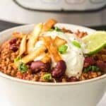 A bowl of Instant Pot Chili with ground meat, kidney beans, tomato sauce, topped with sour cream, tortilla strips, green onions, and a lime wedge. An appliance is visible in the background.