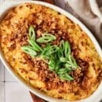 Oval casserole dish filled with Mashed Potato Casserole, topped with crispy fried onions and sliced green onions, placed on a tiled surface next to a beige cloth.
